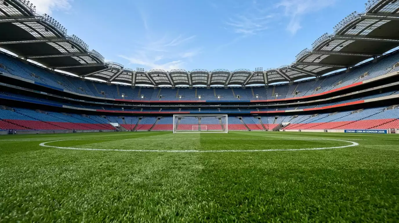 Staidiam sacair mór le páirc glas agus suíocháin fholmha faoi spéir ghorm roimh Chorn an Domhain 2026
